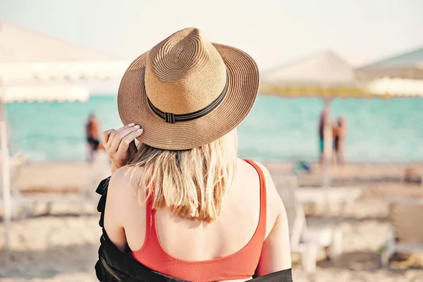 Vacker flicka i en röd bikini och stråhatt på stranden. Back View. Vit sand, blått vatten och parasoller på bakgrunden. Rese-och avkopplings koncept. — Stockfoto
