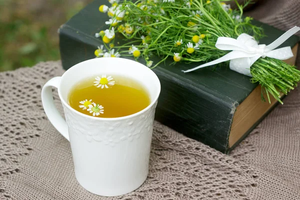 Een kopje van nuttige rustgevende kamille thee, een boeket van chamomiles en een boek op een schommel. — Stockfoto