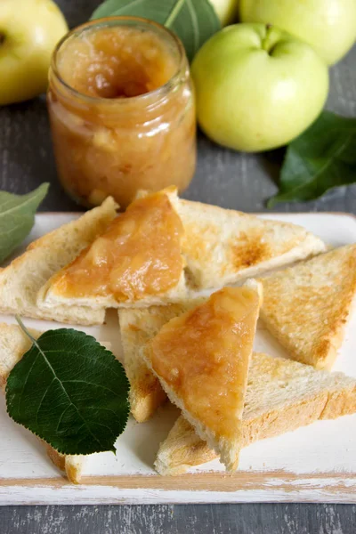 Apple jam in a glass jar with bread toasts and ripe apples on a wooden surface. Rustic style, selective focus.