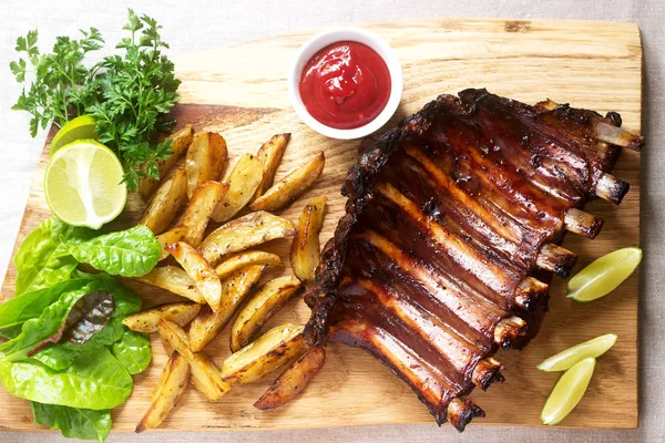 Homemade baked meat ribs served with french fries, herbs, lime and ketchup on a wooden board. Rustic style, selective focus.