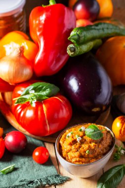 Cam kavanozda patlıcan havyarı ve tahta arka planda sebzeler. Patlıcan, biber, soğan, sarımsak, domates, fesleğen: sebze havyarı için malzemeler. Kırsal biçim, seçici odak.