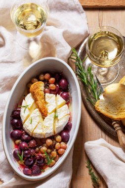 Baked Camembert cheese with grapes, thyme, and rosemary, served with white wine and a baguette. Appetizer on a wooden surface. Rustic style, selective focus.