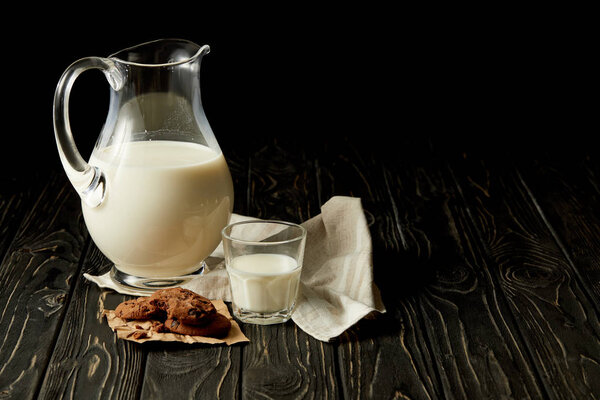 closeup view of fresh milk in jug and glass, chocolate cookies and sackcloth on black background 