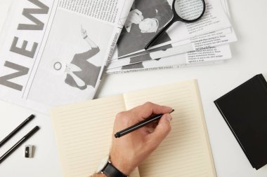 partial top view of person writing in blank notebook, newspapers, magnifying glass, notebook and office supplies on grey clipart
