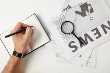 partial top view of person writing in blank notebook and magnifying glass on newspapers on grey clipart
