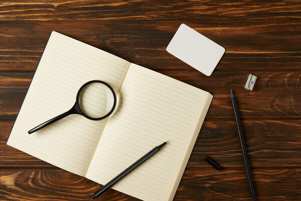 top view of magnifying glass, blank notebook and office supplies on wooden table