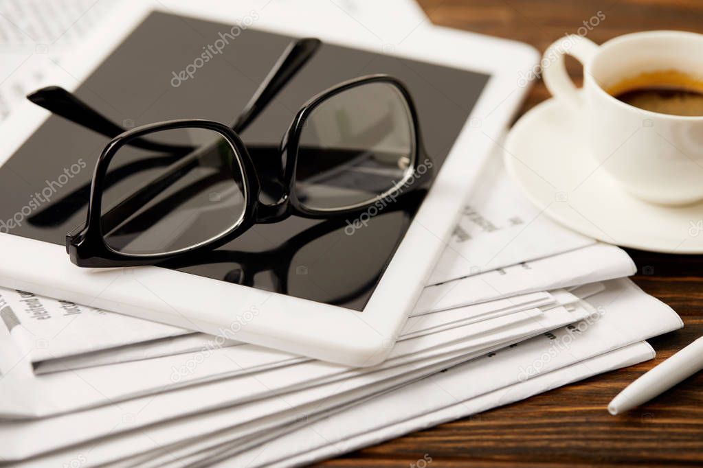 eyeglasses, coffee cup, digital tablet and newspapers on wooden table