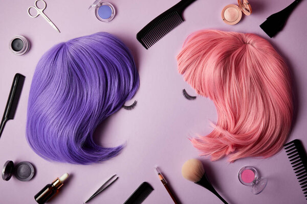 top view of pink and violet wigs, false eyelashes, makeup tools and cosmetics on purple