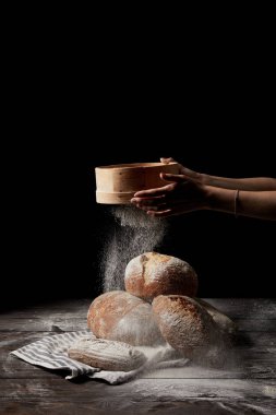 cropped shot of female baker sieving flour over various types of bread on sackcloth isolated on black background  clipart