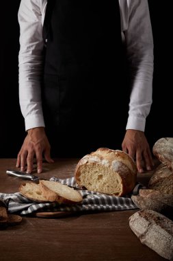 cropped image of male baker in apron standing near table with bread, knife and sackcloth clipart