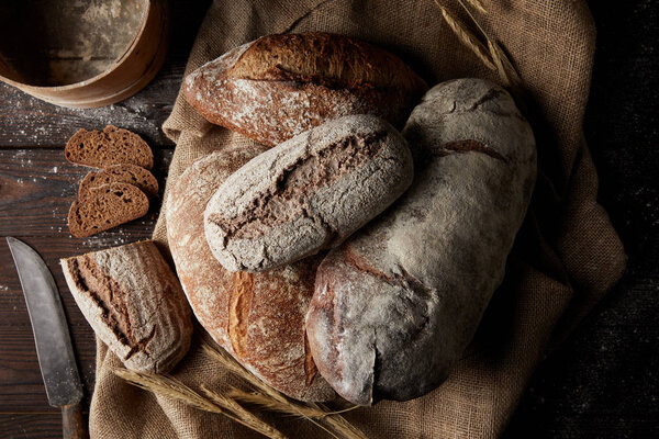 top view of various types of bread, wheat, knife, sieve and sackcloth on wooden table 