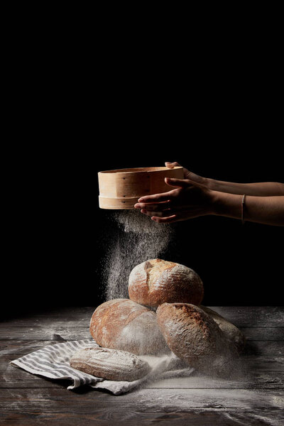 cropped shot of female baker sieving flour over various types of bread on sackcloth isolated on black background 