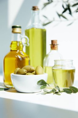 various bottles of aromatic olive oil, jar, bowl with green olives and branches on white table clipart
