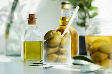 glass with spoon and green olives, jar, various bottles of aromatic olive oil with and branches on white table clipart