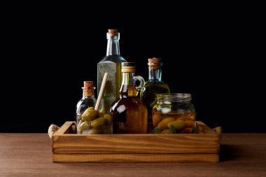 various bottles of aromatic olive oil and jar with green olives on wooden table on black background clipart