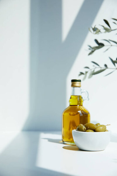 bottle of aromatic olive oil with branches and bowl with green olives on white table