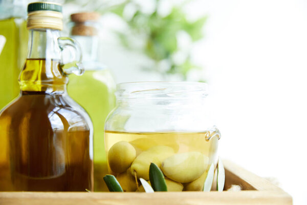 close up shot of jar with green olives, bottles of aromatic olive oil with and branches on wooden tray