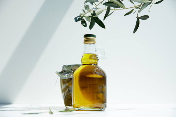 bottle of aromatic olive oil, branches and jar with green olives on white table