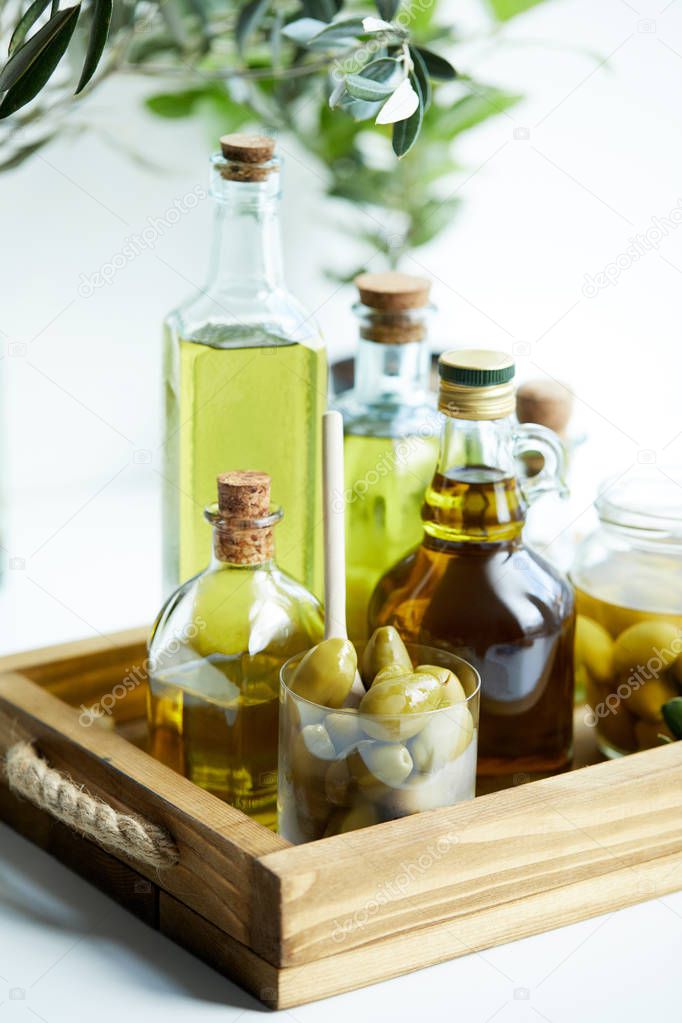 glass with spoon and olives, jar, various bottles of aromatic olive oil with and branches on wooden tray