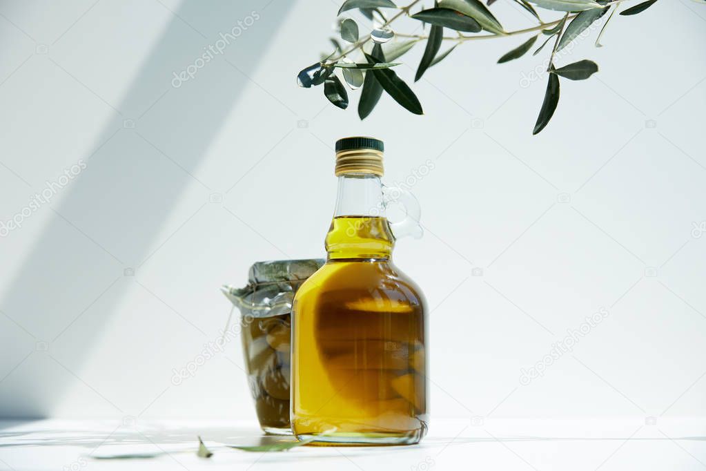 bottle of aromatic olive oil, branches and jar with green olives on white table
