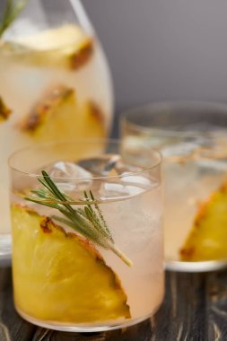selective focus of jug and two glasses of lemonade with pineapple pieces, ice cubes and rosemary on grey wooden tablet clipart