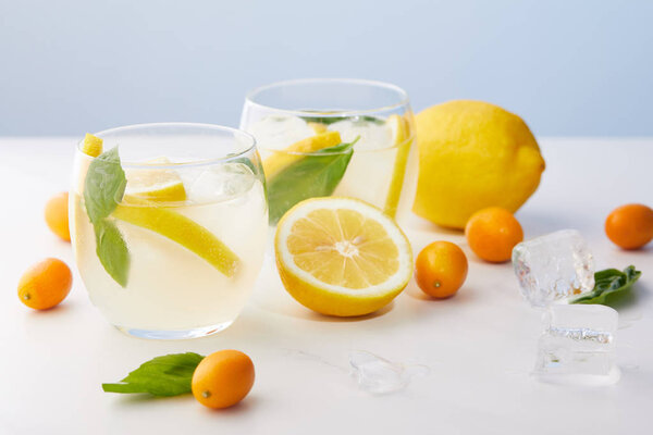два стакана лимонада с мятными листьями, кубиками льда и ломтиками лимона в окружении кумкватов и лимонов на синем фоне
 
