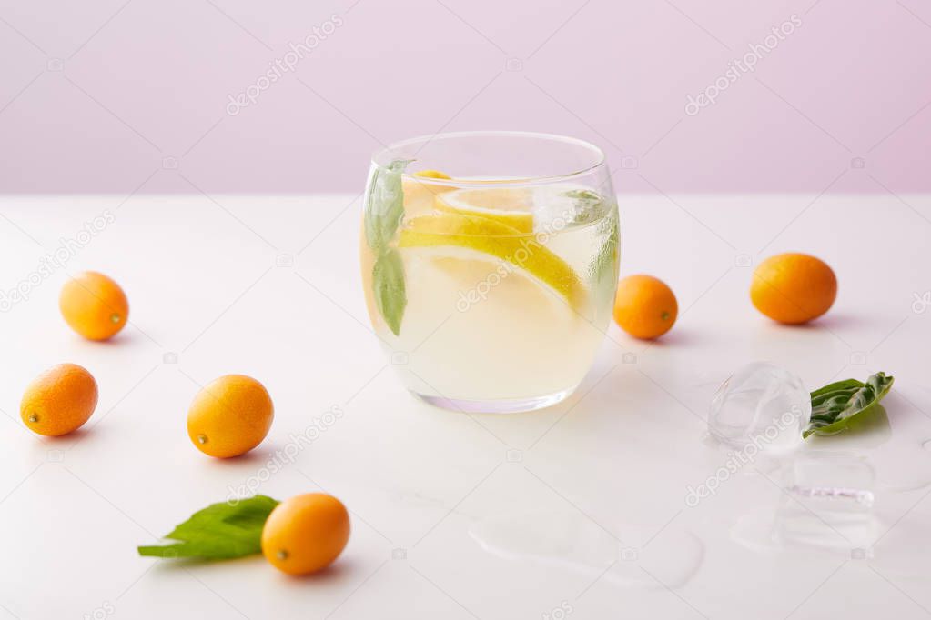 glass of lemonade with mint leaves and lemon slices surrounded by ice cubes, kumquats on purple background 