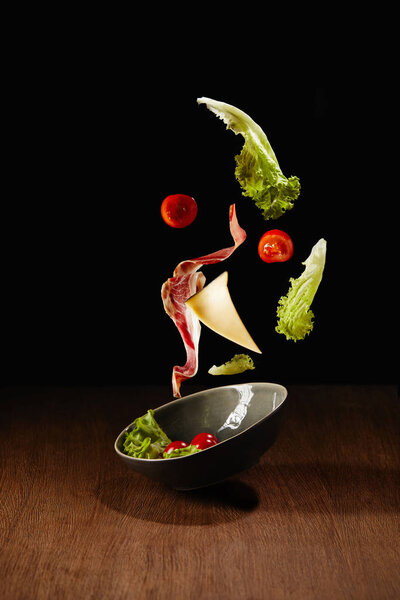 Fresh salad with meat and cheese falling in bowl above wooden table surface