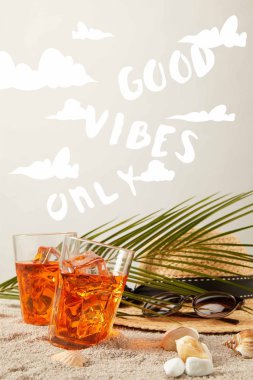 close up view of straw hat, cocktails, seashells, sunglasses and palm leaf on sand on grey backdrop with 