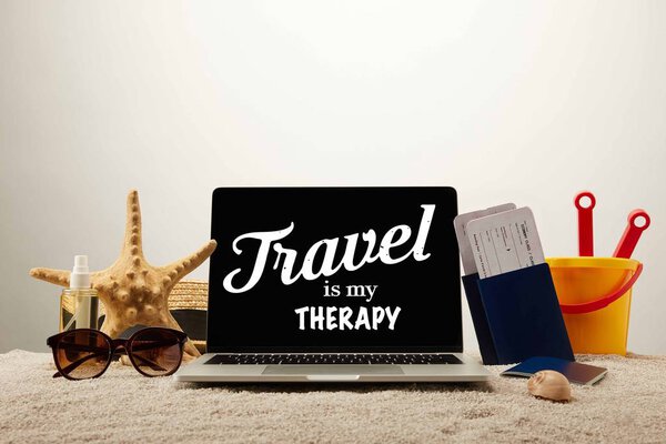 close up view of laptop with "travel is my therapy" lettering, sea star, sunglasses, passports with tickets and toy bucket on sand on grey background
