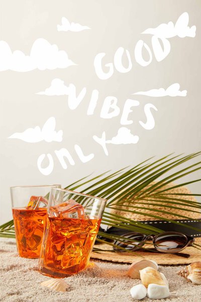 close up view of straw hat, cocktails, seashells, sunglasses and palm leaf on sand on grey backdrop with "good vibes only" sign