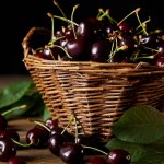 Ripe harvested cherries in rustic basket with leaves on wooden table and on black