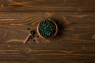 top view of wooden spoon and bowl with pile of spirulina pills on wooden table clipart