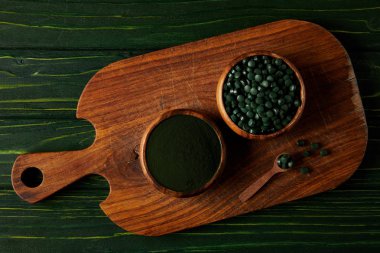 top view of cutting board with wooden spoon, bowls with spirulina powder and spirulina in pills on green wooden table  clipart