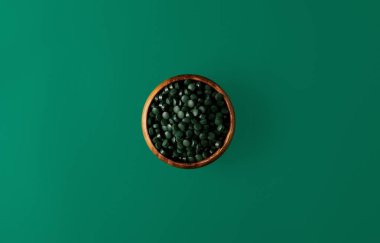 top view of wooden bowl with pile of spirulina pills isolated on green   clipart