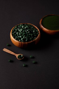 wooden spoon and bowls with ground spirulina and spirulina in pills on grey table  clipart