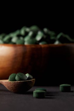 selective focus of wooden spoon and bowl with pile of spirulina pills on black background  clipart
