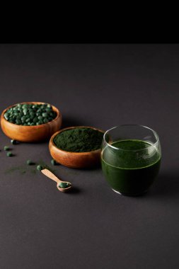 wooden spoon, glass of fresh spirulina smoothie, bowls with spirulina powder and spirulina pills on grey table  clipart