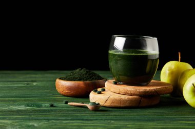 selective focus of wooden slices with fresh spirulina drink in glass, apples, wooden spoon and spirulina powder in wooden bowl on black background on black background  clipart
