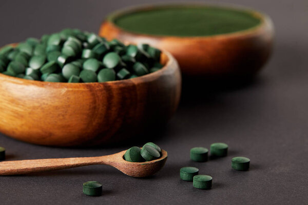 selective focus of wooden spoon and bowls with ground spirulina and spirulina in pills on grey table 