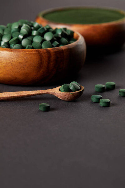 selective focus of wooden spoon, bowls with ground spirulina and spirulina in pills on grey table 