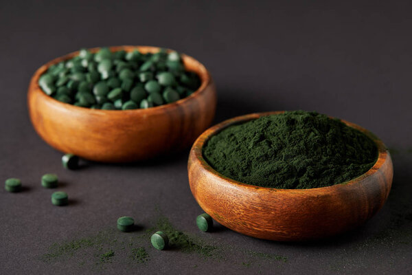 селективный фокус деревянных чаш со спирулиной и спирулиной в таблетках на сером столе
 