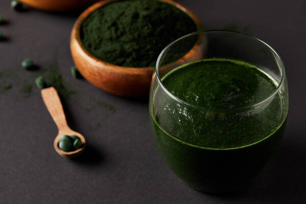 close up view of wooden spoon, glass of smoothie from spirulina, bowls with spirulina powder and spirulina pills on grey table 