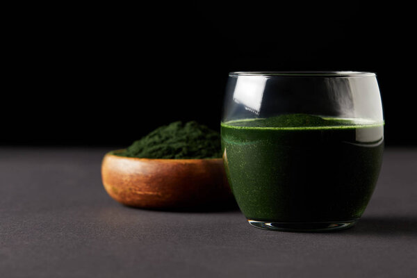 close up view of fresh spirulina drink in glass and spirulina powder in wooden bowl on black background 