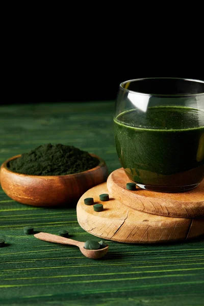 Cam, tahta kaşık ve spirulina tozu yeşil ahşap masa üzerinde siyah arka plan üzerine ahşap kase taze spirulina ile Ahşap dilimlerin içki 