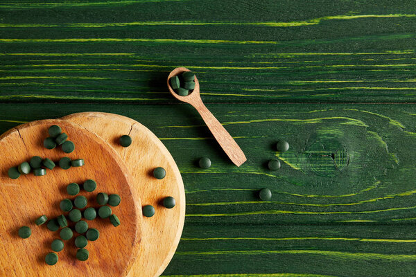 top view of wooden cutting boards, spoon and spirulina pills on green wooden table