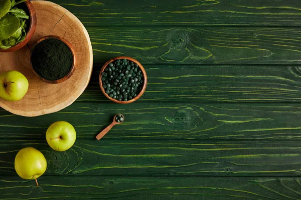 yeşil ahşap masa üzerinde kesme tahtası, ahşap tabak, kaşık, elma, yaprakları, spirulina tozu ve spirulina hapları üstten görünüm 