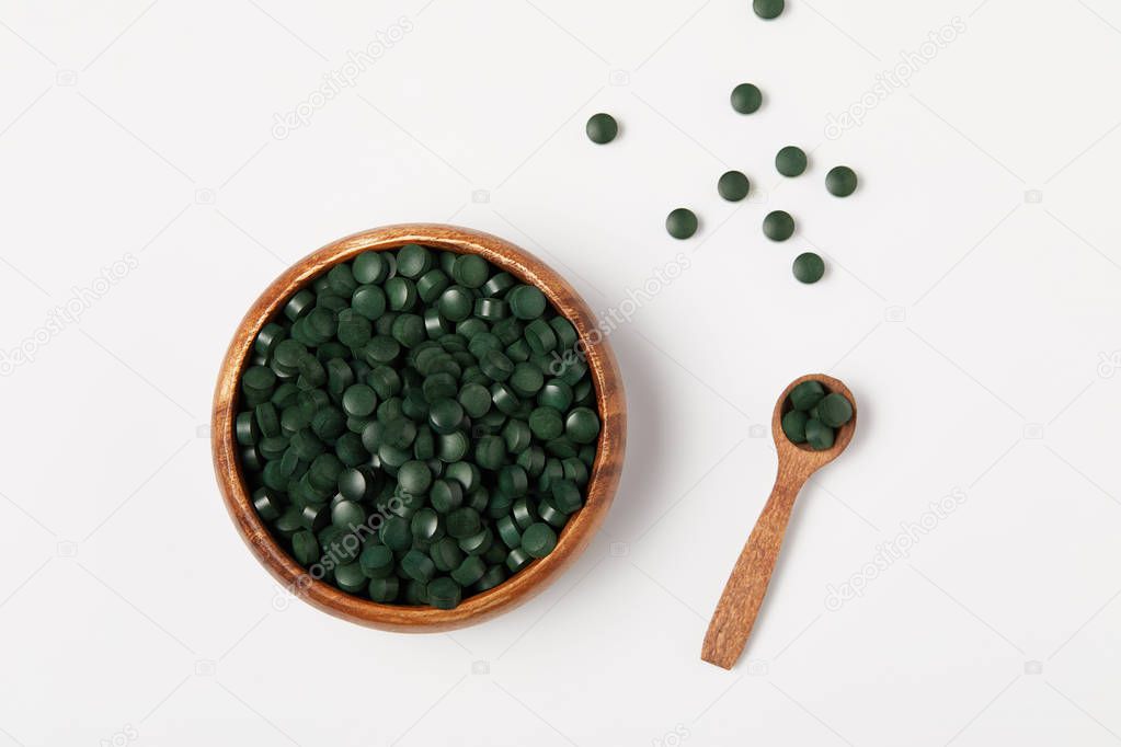 elevated view of wooden spoon and bowl with pile of spirulina pills on grey table 