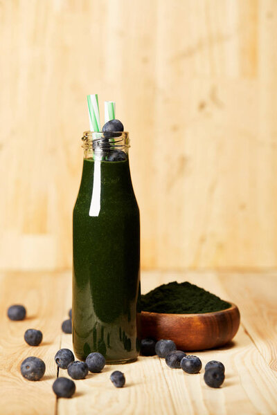 bowl with spiruluna powder, bottle of spirulina smoothie with blueberries and drinking straw on wooden table