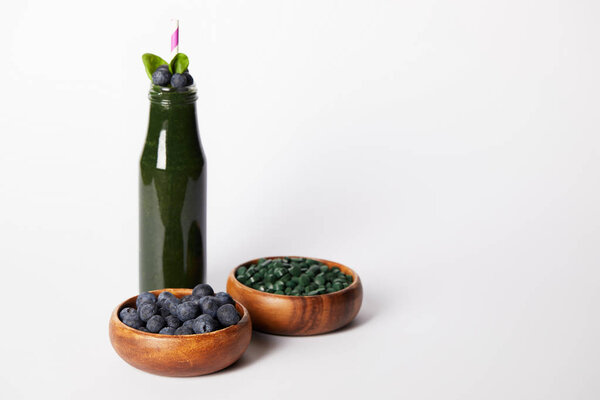 bottle of spirulina smoothie with mint leaves and drinking straw, bowls with blueberries and spirulina pills on grey background 